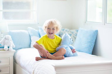 Beyaz güneşli bir odada pencereli bir çocuk yatakta oynuyor. Çocuk odası ve iç tasarım. Erkek bebek evde. Çocuk yuvası için yatak ve tekstil. Oyuncağı ve kitabı olan bir çocuk. Uyku ve uyku zamanı..
