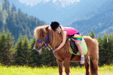 Alp dağlarında midilliye binen çocuklar. Avusturya 'daki at çiftliğinde aile tatili, Tirol. Çocuklar ata biner. Çocuk hayvanla ilgileniyor. Çocuk ve evcil hayvan. Çiçek açan çayırdaki küçük kız.