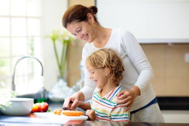 Anne ve çocuk beyaz mutfakta yemek yapıyorlar. Aile evde. Anne ve küçük çocuk sağlıklı öğle yemeği için taze sebze doğradılar. Ebeveynler ve çocuklar kaynaşıyor. Genç kadın ve çocuğu yemek pişiriyor..
