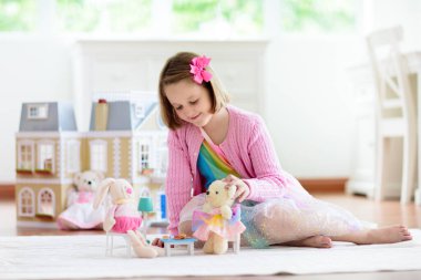 Beyaz güneşli yatak odasında oyuncak bebek eviyle oynayan küçük bir kız. Oyuncaklı çocuk. Küçük çocuklar için rol oyunu. Oyuncak ayılı çocuk. Çocuklar oyuncak hayvan ve bebeklerle çay partisi yapıyorlar. Çocuk odası.