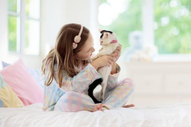 Beyaz yatak odasında bebek kediyle oynayan bir çocuk. Beyaz kediciği tutan çocuk. Evde sevimli bir hayvanı olan pijamalı küçük bir kız. Çocuklar kedilerle oynar. Çocuklar ve evcil hayvanlar.