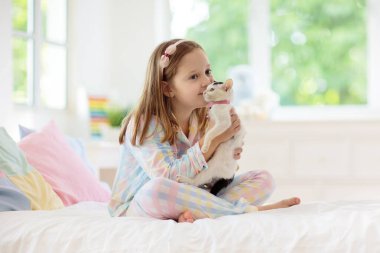 Beyaz yatak odasında bebek kediyle oynayan bir çocuk. Beyaz kediciği tutan çocuk. Evde sevimli bir hayvanı olan pijamalı küçük bir kız. Çocuklar kedilerle oynar. Çocuklar ve evcil hayvanlar.
