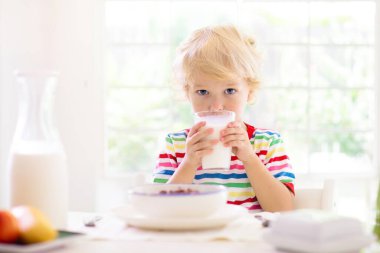 Çocuk kahvaltı yapıyor. Çocuk süt içiyor ve meyveli mısır gevreği yiyor. Pencerenin önündeki mutfakta beyaz yemek masasında oturan küçük çocuk. Çocuklar güneşli sabahlarda yemek yerler. Genç çocuklar için sağlıklı dengeli beslenme.