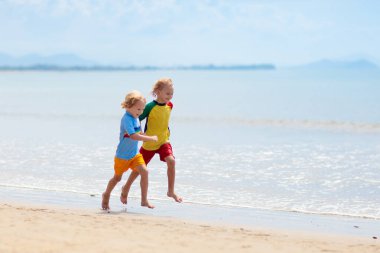 Tropik plajda oynayan çocuklar. Çocuklar yaz tatilinde yüzer ve denizde oynarlar. Kum ve su eğlencesi, küçük çocuklar için güneş koruması. Okyanus kıyısında koşan ve zıplayan küçük çocuk.