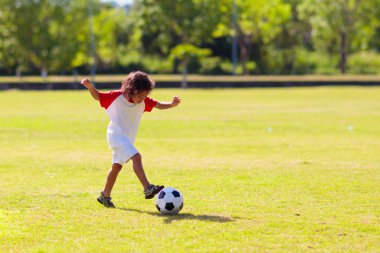 Futbol oynayan tatlı kıvırcık çocuk. Çocuklar açık havada oynarlar. Sevimli küçük atlet topa vuruyor. Yazın açık hava eğlencesi. Bebek topun peşinden koşuyor. Aktif çocuklar için spor. Çocuk egzersizi.