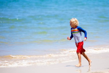 Tropik plajda oynayan çocuklar. Çocuklar yaz tatilinde yüzer ve denizde oynarlar. Kum ve su eğlencesi, küçük çocuklar için güneş koruması. Okyanus kıyısında koşan ve zıplayan küçük çocuk.