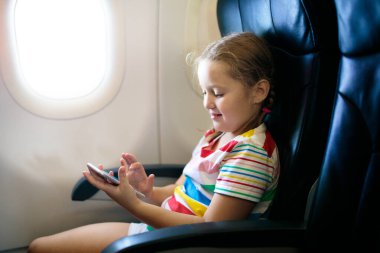 Çocuk uçak. Pencere kenarı düzlemde hava içinde hareket eden telefon ile evlat. Çocuklar için uçuş eğlence. Küçük çocuklarla seyahat. Çocuklar sinek ve seyahat. Aile tatili. Smartphone ile oynayan kız.