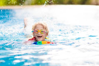 Çocuk tropikal tatil köyü havuzunda yüzmeyi öğreniyor. Çocuklar yüzmeyi öğrenir. Küçük çocuklar için egzersiz ve eğitim. Spor kulübünde renkli sörf tahtası olan küçük bir çocuk. Yüzen bebek ya da bebek.