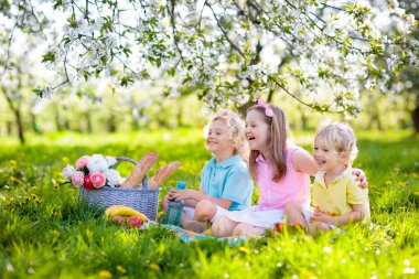 Bahar Parkı 'nda çiçek açan kiraz ağaçlarıyla aile pikniği. Çocuklar dışarıda meyve ve ekmek yiyorlar. Çiçekli elma bahçesinde piknik sepetiyle battaniyenin üzerinde oturuyorlar. Çocuklar için sağlıklı beslenme.