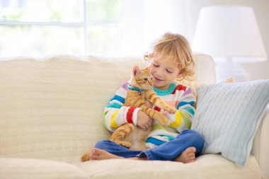 Çocuk kediyle oynuyor. Kedi yavrusu tutan çocuk. Küçük çocuk evdeki güneşli oturma odasında kanepede oturan sevimli hayvanlara sarılıyor. Çocuklar evcil hayvanlarla oynar. Çocuklar ve evcil hayvanlar.