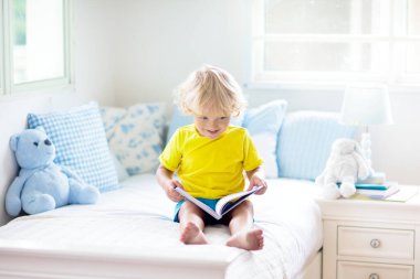 Beyaz güneşli bir odada pencereli bir çocuk yatakta oynuyor. Çocuk odası ve iç tasarım. Erkek bebek evde. Çocuk yuvası için yatak ve tekstil. Oyuncağı ve kitabı olan bir çocuk. Uyku ve uyku zamanı..