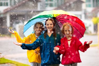 Çocuklar sonbahar yağmurunda oynarlar. Çocuklar soğuk yağmurlu havada şehir caddesinde yürürler. Açık havada su geçirmez giysi. Renkli şemsiyeli ve yağmurluklu kız ve erkek. Açık hava eğlencesi.