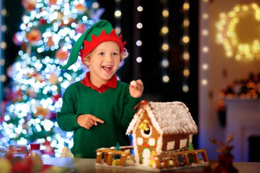 Çocuklar Noel kurabiyesi evi pişiriyor. Çocuklar kış tatilini evde kutluyorlar. Şömine ve ağaçla dekore edilmiş oturma odası. Aile aktivitesi. Küçük çocuk kurabiye yapıyor..