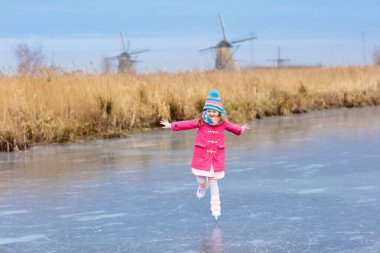 Çocuk buz dondurulmuş kanalda rüzgar değirmenleri ve Hollanda'da kar buz pateni. Hollanda soğuk kış gününde doğal buz paten olan küçük kız. Çocukların buz pateni karlı Hollanda Yeldeğirmeni peyzaj.