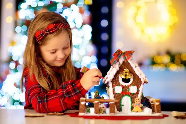 Çocuklar Noel kurabiyesi evi pişiriyor. Çocuklar kış tatilini evde kutluyorlar. Şömine ve ağaçla dekore edilmiş oturma odası. Aile aktivitesi. Küçük kız kurabiye yapıyor..