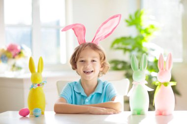 Çocuklar paskalya yumurtalarını boyuyor. Tavşan kulaklı çocuklar Paskalya avı için renkli yumurta boyuyorlar. Bahar tatili kutlaması için çiçek, sepet ve tavşanlı ev dekorasyonu. Küçük çocuk evi dekore ediyor..