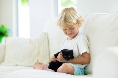 Çocuk yavru kediyle oynuyor. Siyah kediciği tutan çocuk. Küçük çocuk evdeki güneşli oturma odasında beyaz koltukta oturan sevimli evcil hayvana sarılıyor. Çocuklar evcil hayvanlarla oynar. Çocuklar ve evcil hayvanlar.