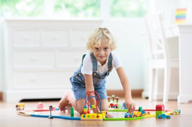 Çocuklar ahşap demiryolu ile oynarlar. Oyuncak trenli çocuk. Küçük çocuklar için eğitici oyuncaklar. Küçük bir çocuk, beyaz zeminde ya da anaokulunda demiryolu inşa ediyor. Araba ve motor çalan sevimli bir çocuk..