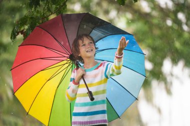 Sonbahar yağmurunda oynayan çocuk. Parktaki çamurlu su birikintisine atlayan çocuk. Şiddetli sonbahar yağmuru altında renkli şemsiyeli küçük bir çocuk. Fırtınalı yağmurlu hava eğlencesi.