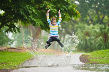 Sonbahar yağmurunda oynayan çocuk. Parktaki çamurlu su birikintisine atlayan çocuk. Şiddetli sonbahar yağmuru altında renkli şemsiyeli küçük bir çocuk. Fırtınalı yağmurlu hava eğlencesi.