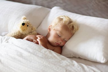 Bebek beyaz yatakta uyuyor. Sağlıklı çocuklar uyur. Küçük çocuk ebeveyn yatağında kestiriyor. Bebek için güneşli bir kreş. Küçük çocuk için yatak. Uyku vakti. Güneşli bir sabahta evde yeni yürümeye başlayan çocuk..