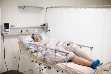 Modern hastanenin ameliyat sonrası bakım odasında iyileşmekte olan genç bir hasta. Diz travması sonrası alçı ve nabız ölçer olan kadın ortopedi kliniğinin dinlenme odasında izleniyor..