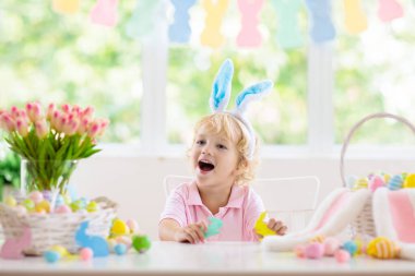 Çocuklar paskalya yumurtalarını boyuyor. Tavşan kulaklı çocuklar Paskalya avı için renkli yumurta boyuyorlar. Bahar tatili kutlaması için çiçek, sepet ve tavşanlı ev dekorasyonu. Küçük bebek evi dekore ediyor..