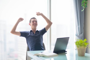 Şehir manzaralı büyük pencerede dizüstü bilgisayarda çalışan bir adam. Merkez ofis. Bilgisayarlı genç serbest çalışan bir erkek. Öğrenci çevrimiçi çalışıyor. Cep telefonu ve bilgisayarı olan adam. Evden çalış..