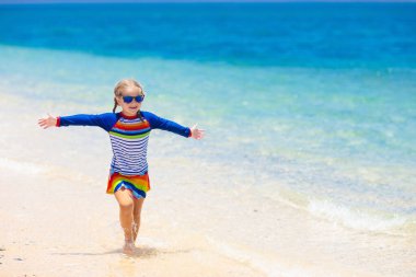 Tropik plajda oynayan çocuklar. Çocuklar yaz tatilinde yüzer ve denizde oynarlar. Kum ve su eğlencesi, küçük çocuklar için güneş koruması. Okyanus kıyısında koşup zıplayan küçük kız.