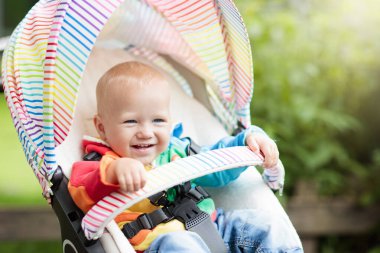 Beyaz kazaklı bebek, parkta beyaz bir bebek arabasında oturuyor. Renkli gökkuşağı arabalı çocuk. Tekerlekli sandalyedeki küçük çocuk. Küçük çocuklarla seyahat etmek. Bebekli aile için ulaşım.