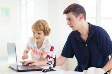Mikroskoplu okul çocukları. Fen dersi. Dizüstü bilgisayarla biyoloji ya da kimya deneyi yapan çocuklar. Çevrimiçi öğrenme. İlkokul çocuğuna uzaktan eğitim. Çocuk konuya bakıyor..