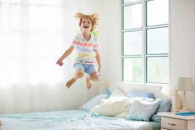 Büyük pencereli beyaz yatak odasında yatağa atlayan bir çocuk. Çocuklar evde oynuyor. Ebeveyn yatağında eğlenceli bir atlayış. Oda ve iç tasarım. Evdeki çocuk. Güneşli bir sabah, hafta sonu aile dinlenme zamanı..