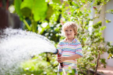 Bahçede çiçekleri ve bitkileri sulayan çocuk. Güneşli bahçesinde su hortumu olan bir çocuk. Küçük çocuk bahçeyle uğraşıyor. Evde yaz eğlencesi. Baharda aile aktivitesi. Çocuklar ebeveynlere yardım.