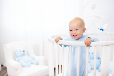 Komik küçük bebek yuvarlak modern yatakta hareket eden pencere ile beyaz kreş ile ayakta. Mavi pijama ile oyuncak ayı oyuncak bebek boy. Genç çocuk için çocuk odası iç. Çocuklar uyku giyim ve yatak takımları.