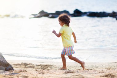 Tropik plajda oynayan tatlı kıvırcık çocuk. Çocuklar deniz kıyısında oynuyorlar. Su ve kum eğlencesi. Yaz tatili. Çocuklarla seyahat et. Çocuklu bir okyanus tatili. Sahilde gün batımı yürüyüşü.