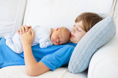 Uyuyan bebek kardeşi tutan mutlu gülen çocuk. Büyük yaş farkı ile kardeş. Çocuklar evde beyaz kanepede oynuyor. Yeni doğan çocuk kestiriyor. Bağ çocuklar. Aile sevgisi. 