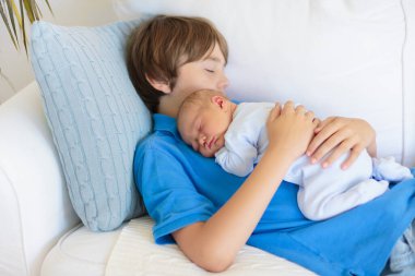 Uyuyan bebek kardeşi tutan mutlu gülen çocuk. Büyük yaş farkı ile kardeş. Çocuklar evde beyaz kanepede oynuyor. Yeni doğan çocuk kestiriyor. Bağ çocuklar. Aile sevgisi. 