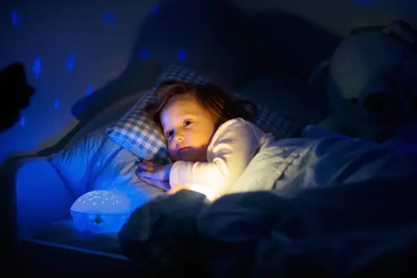 stock image Little girl reading a book in bed. Dark bedroom with night light projecting stars on room ceiling. Kids nursery and bedding. Children read before bedtime. Toddler child playing with lamp and bear toy.