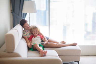 Çocuk ve anne güneşli odada bir kitap okudu. Küçük çocuk ve anne, şehir manzaralı büyük bir penceredeki kanepede masal okuyorlar. Modern gökdelende sıcak bir sabah. Aile zamanı..