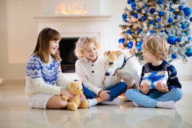 Evde Noel. Çocuklar ve köpek yılbaşı ağacının altında. Küçük oğlan ve kız Noel Baba şapkalı evcil hayvana sarılıp Noel hediyelerini açıyor. Çocuklar hayvanlarla oynar. Kış bayramı kutlaması. Mavi ve beyaz tema.