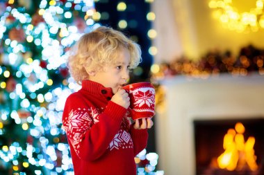 Çocuk evde Noel ağacı sıcak çikolata içme. Süslü içecek kupa. Sıcak kakao kırmızı Kupası tutan Xmas süsleme ile örme kazak içindeki küçük çocuk. Çocuklar ailesiyle kutlamak kış tatili.