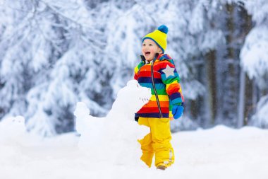 Karlı kış parkta kardan adam yapma çocuk. Çocuk karda oyun. Renkli ceket ve sonra kar yağışı kar dinozor kış bahçesinde bina şapka erkek bebek. Soğuk kış günü açık eğlence.