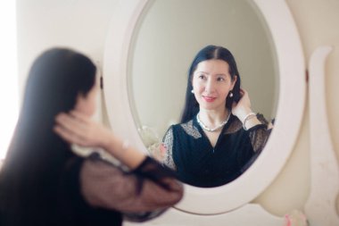 Genç Asyalı kadın aynaya bakıyor. Cilt bakımı rutini. Randevu gecesi ya da hafta sonu sabahında krem süren gecelikli güzel bir kadın. Deri temizleme ve tedavi. Mücevher ve inciler.. 