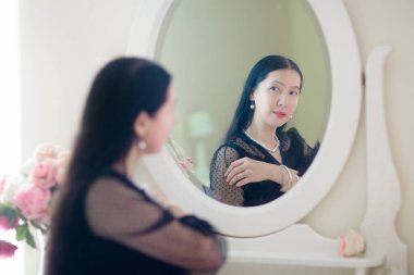 Genç Asyalı kadın aynaya bakıyor. Cilt bakımı rutini. Randevu gecesi ya da hafta sonu sabahında krem süren gecelikli güzel bir kadın. Deri temizleme ve tedavi. Mücevher ve inciler.. 