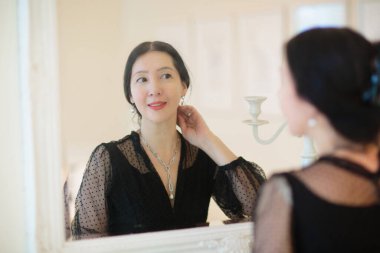 Genç Asyalı kadın aynaya bakıyor. Cilt bakımı rutini. Randevu gecesi ya da hafta sonu sabahında krem süren gecelikli güzel bir kadın. Deri temizleme ve tedavi. Mücevher ve inciler.. 