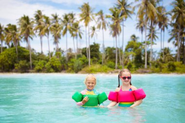 Hindistan cevizi palmiyeli tropik plajdaki çocuklar. Çocuklar için yüzme yardımları. Renkli kol kanatlarıyla yüzen kız ve erkek. Su oyuncakları. Bebek yüzmeyi öğreniyor. Egzotik adada aile deniz tatili.