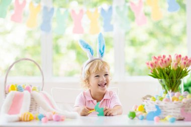 Çocuklar paskalya yumurtalarını boyuyor. Tavşan kulaklı çocuklar Paskalya avı için renkli yumurta boyuyorlar. Bahar tatili kutlaması için çiçek, sepet ve tavşanlı ev dekorasyonu. Küçük bebek evi dekore ediyor..