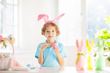 Çocuklar paskalya yumurtalarını boyuyor. Tavşan kulaklı çocuklar Paskalya avı için renkli yumurta boyuyorlar. Bahar tatili kutlaması için çiçek, sepet ve tavşanlı ev dekorasyonu. Küçük çocuk evi dekore ediyor..