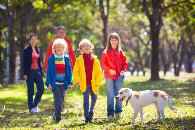 Sonbahar parkında aile yürüyüşü. Çocuklar güneşli ormanda oynarlar. Açık hava eğlencesi. Anne, baba ve çocuklar güneşli sonbahar gününde köpeği gezdirirler. Yağmurluk giyen kız ve erkek. Ailesi oğlu ve kızı var..