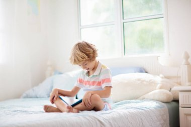 Çocuklar yatakta kitap okuyor. Çocuklar için yatak odası. Küçük çocuk beyaz yatakta renkli yataklı kitaplar okuyor. Çocuk yuvası. Çocuk içeride oynuyor. Şekerleme ve uyku zamanı. Ödev yapan öğrenci.